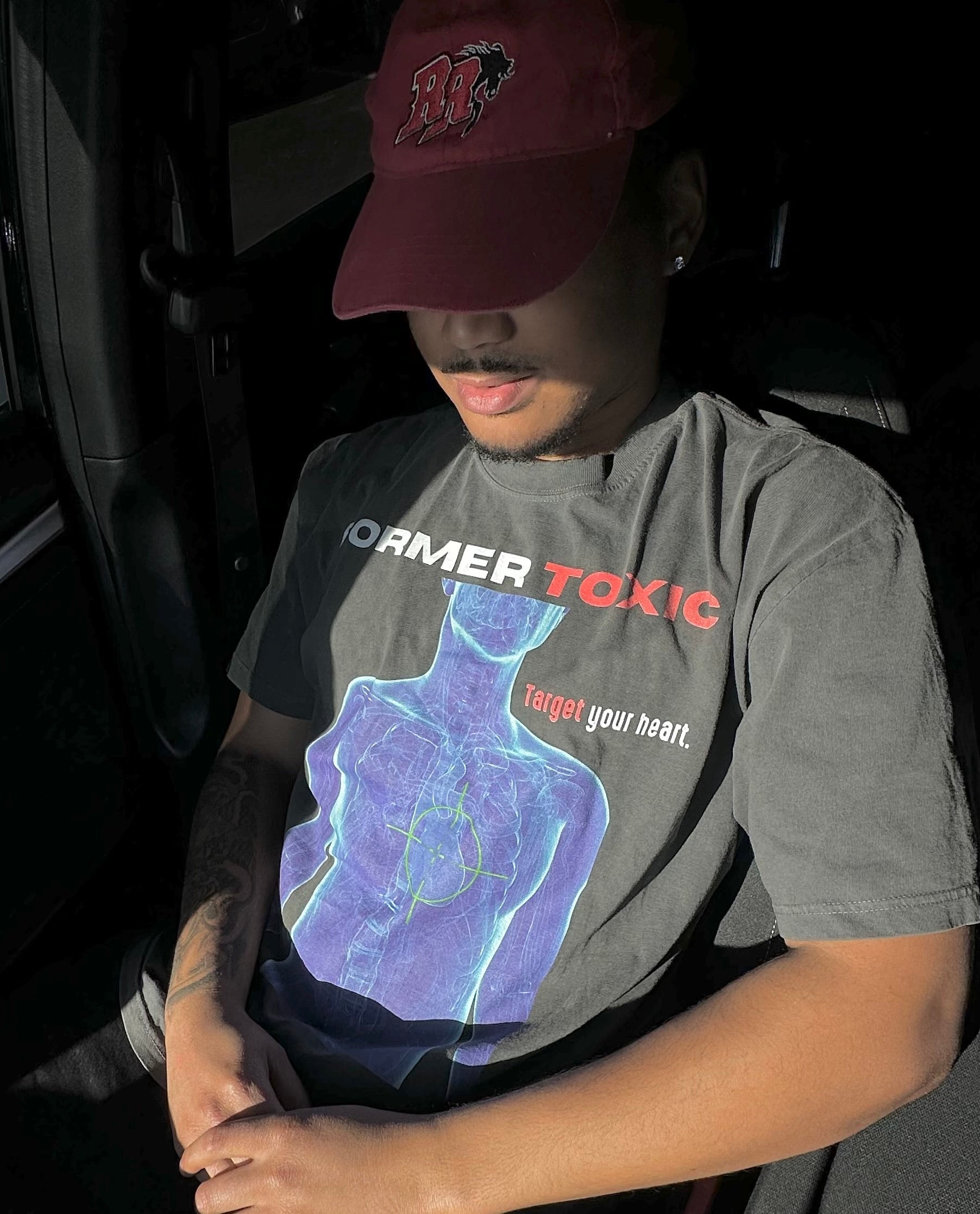 Model wearing charcoal Heat Scan Targeted Heart Tee featuring blue X-ray anatomy graphic and neon heart target while sitting in car.