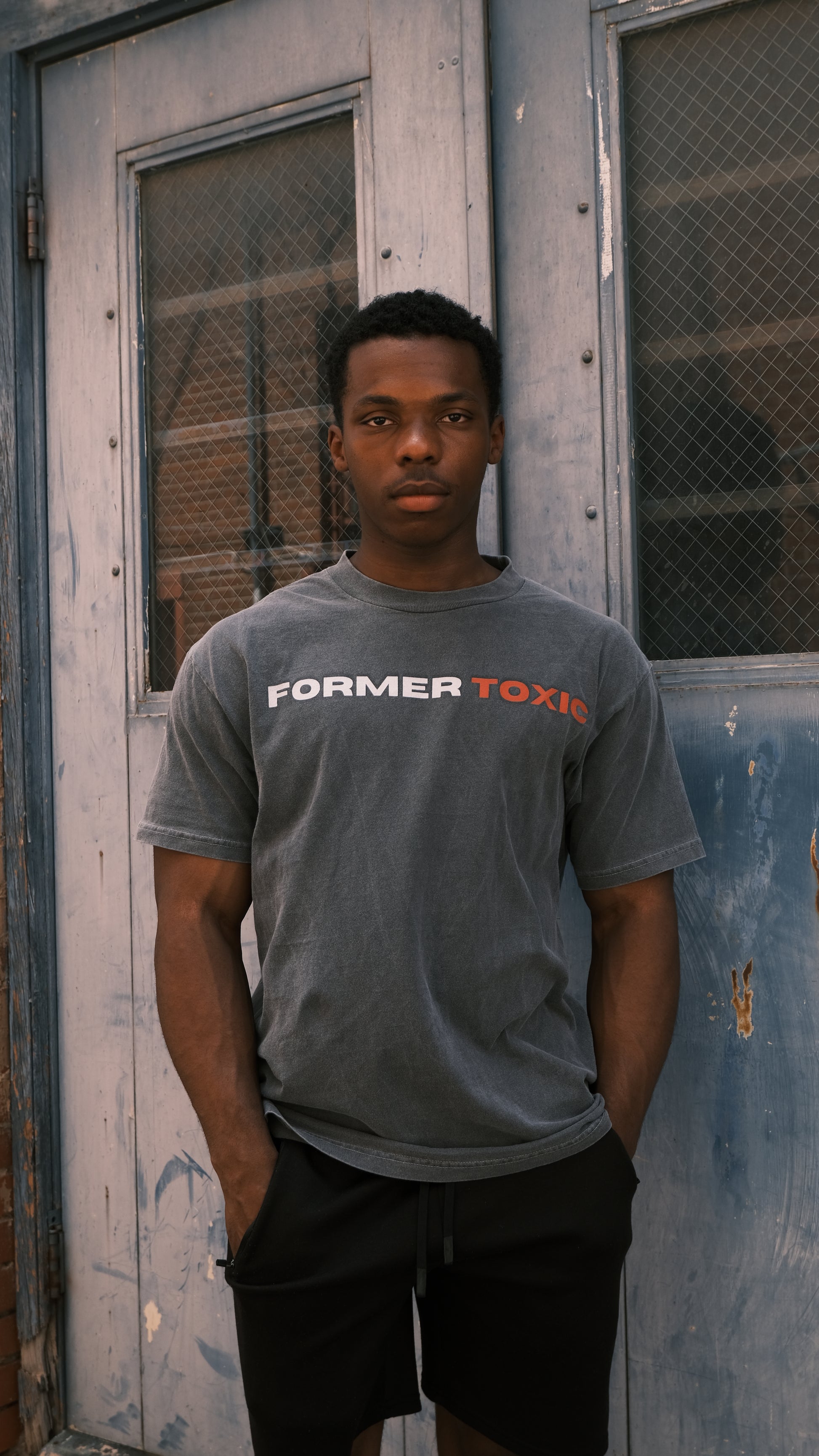 Model wearing washed charcoal Former Toxic Let Love Destroy You tee with bold black and red logo, standing in front of distressed blue door