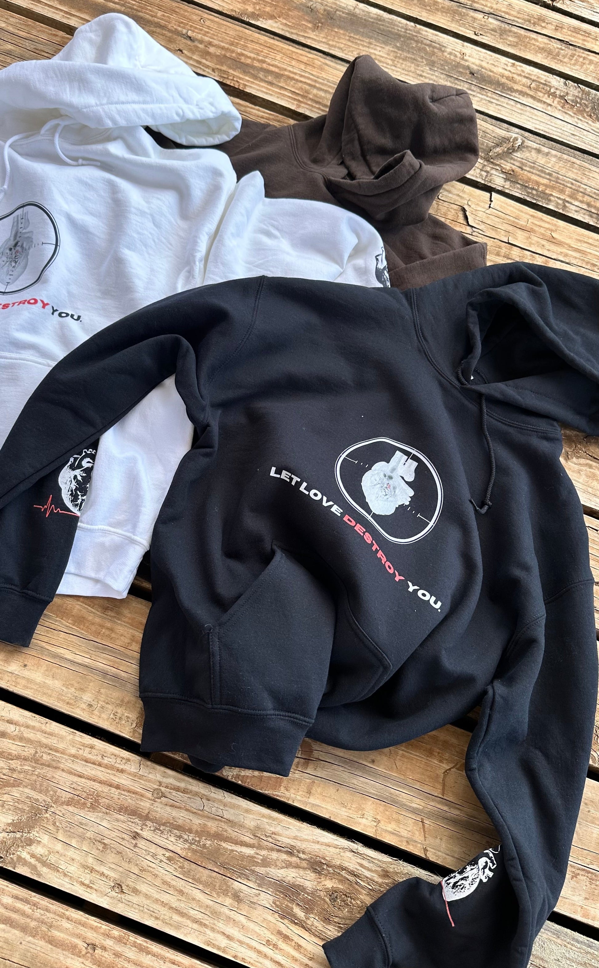Let Love Destroy You hoodies in black, white, and brown displayed on wooden surface showing heart target graphic.