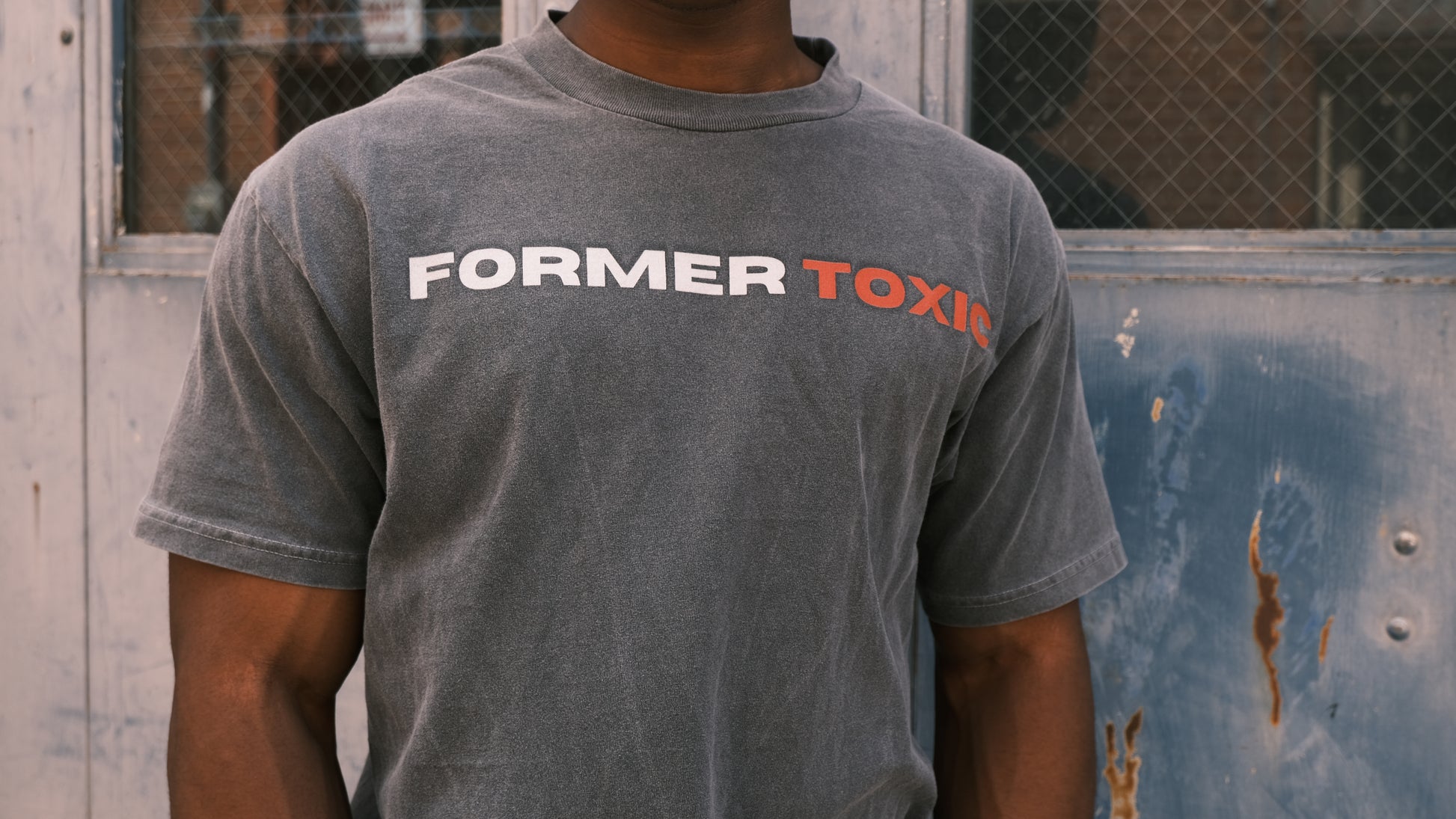 Up close shot of model wearing washed charcoal Former Toxic Let Love Destroy You tee with bold black and red logo, standing in front of distressed blue door