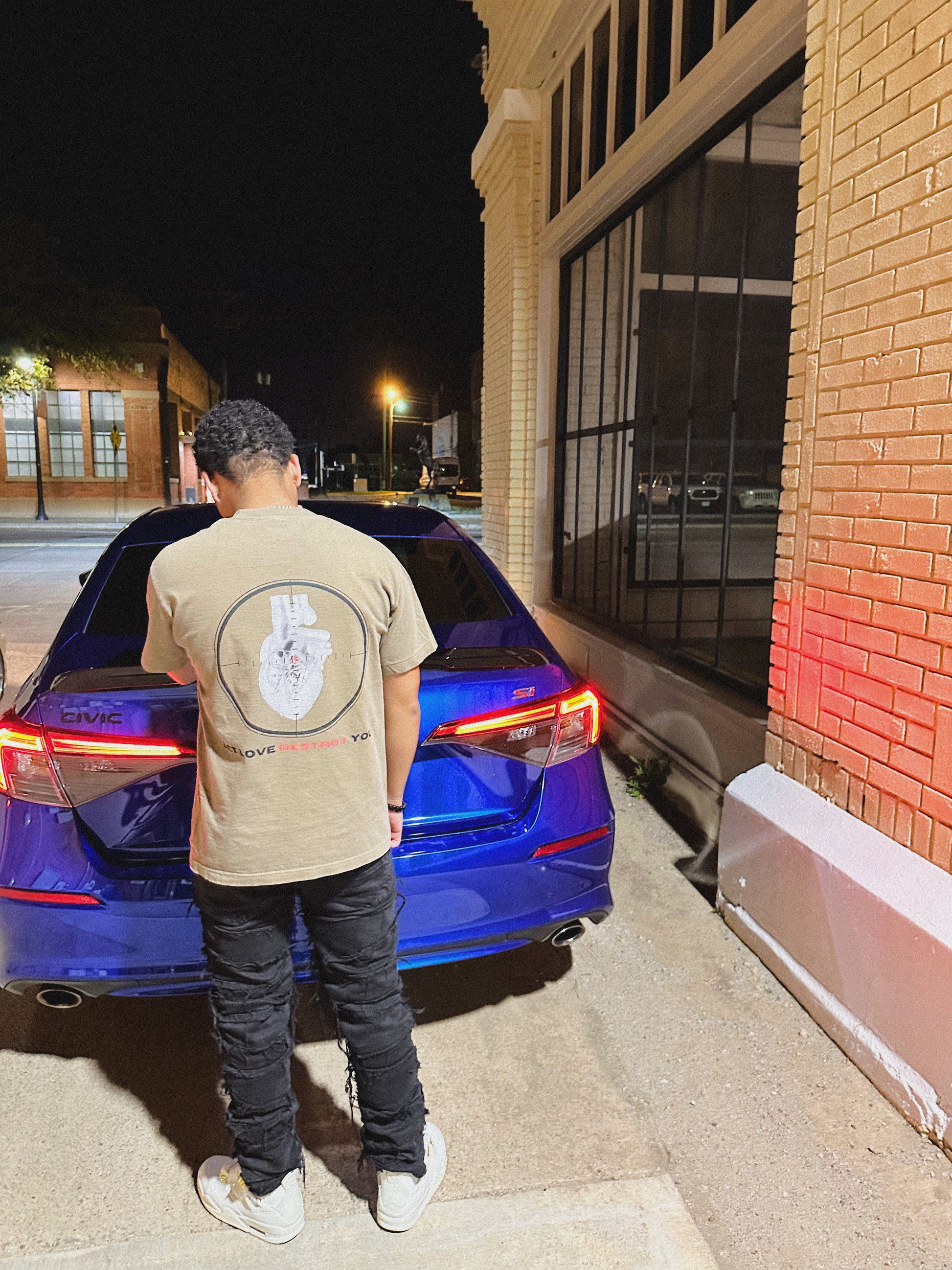Back view of tan Let Love Destroy You tee featuring heart-in-crosshair graphic, photographed outdoors beside blue car at night.