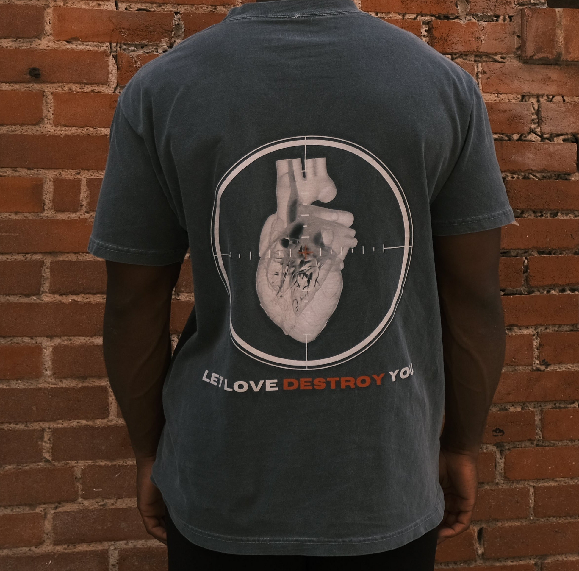 Model wearing washed charcoal Let Love Destroy You tee showing anatomical heart graphic with target design against brick wall.