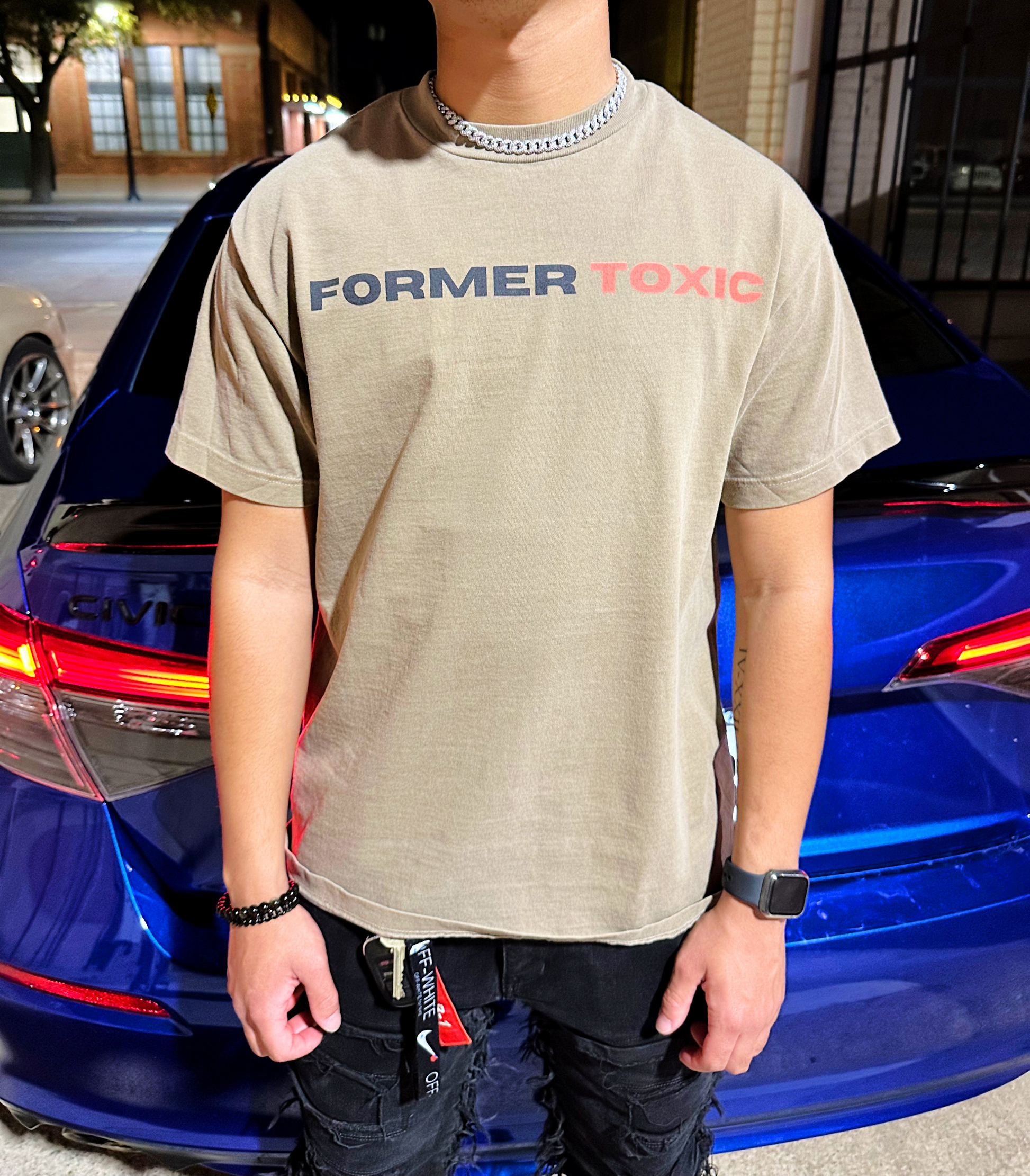 Model wearing tan Former Toxic Let Love Destroy You tee with black and red logo, standing in front of blue car at night.